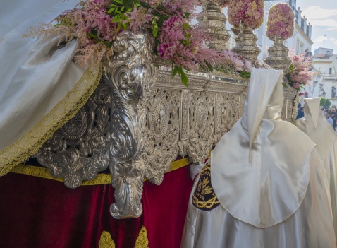 A Semana Santa throne moves through the narrow streets, carried by costaleros beneath its weight; silver engravings gleam beside fresh flowers adorning its base, while Nazarenos in satin robes walk in solemn procession © Michelle Chaplow