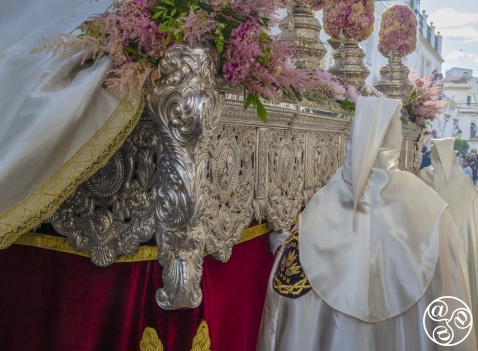 A Semana Santa throne moves through the narrow streets, carried by costaleros beneath its weight; silver engravings gleam beside fresh flowers adorning its base, while Nazarenos in satin robes walk in solemn procession © Michelle Chaplow